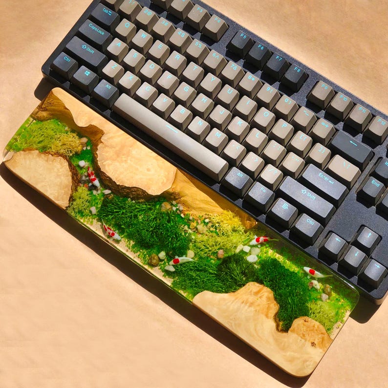 Puede incluir: Un reposamu&ntilde;ecas de madera para teclado con una incrustaci&oacute;n de resina que representa un paisaje de musgo verde y peces rojos y blancos. El reposamu&ntilde;ecas se coloca delante de un teclado negro con teclas beige.