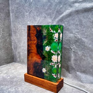May include: A rectangular lamp with a wooden base. The lamp's body features a wood grain design on one side and a green resin design with koi fish, lily pads, and flowers on the other. A white power cord extends from the base.