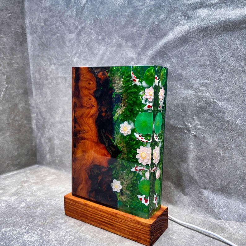 May include: A rectangular lamp with a wooden base. The lamp's body features a wood grain design on one side and a green resin design with koi fish, lily pads, and flowers on the other. A white power cord extends from the base.