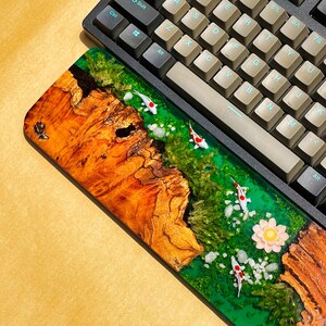 May include: A custom keyboard wrist rest featuring a wood and resin design. The wrist rest has a vibrant green river scene with miniature koi fish, white stones, and a pink flower. The wood has a rich, natural grain.