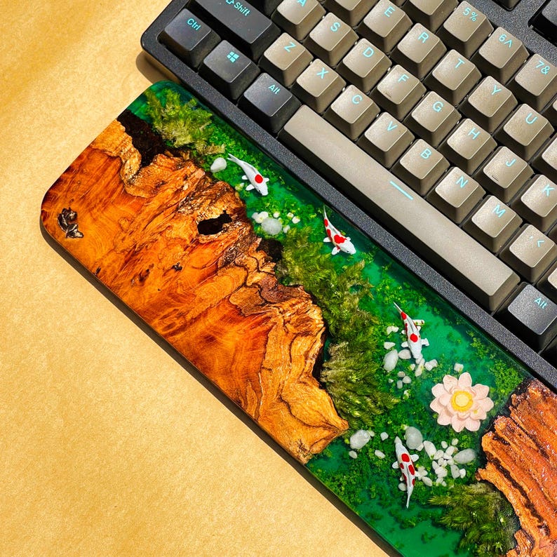 May include: A custom keyboard wrist rest featuring a wood and resin design. The wrist rest has a vibrant green river scene with miniature koi fish, white stones, and a pink flower. The wood has a rich, natural grain.