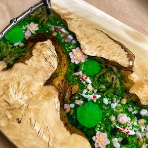Op de afbeelding: Een houten serveerblad met een riviersc&egrave;ne. De rivier is gevuld met groen mos, wit en rode koi-karpers, roze bloemen en groene waterlelies. De lade heeft een lichtbruine houtnerf.