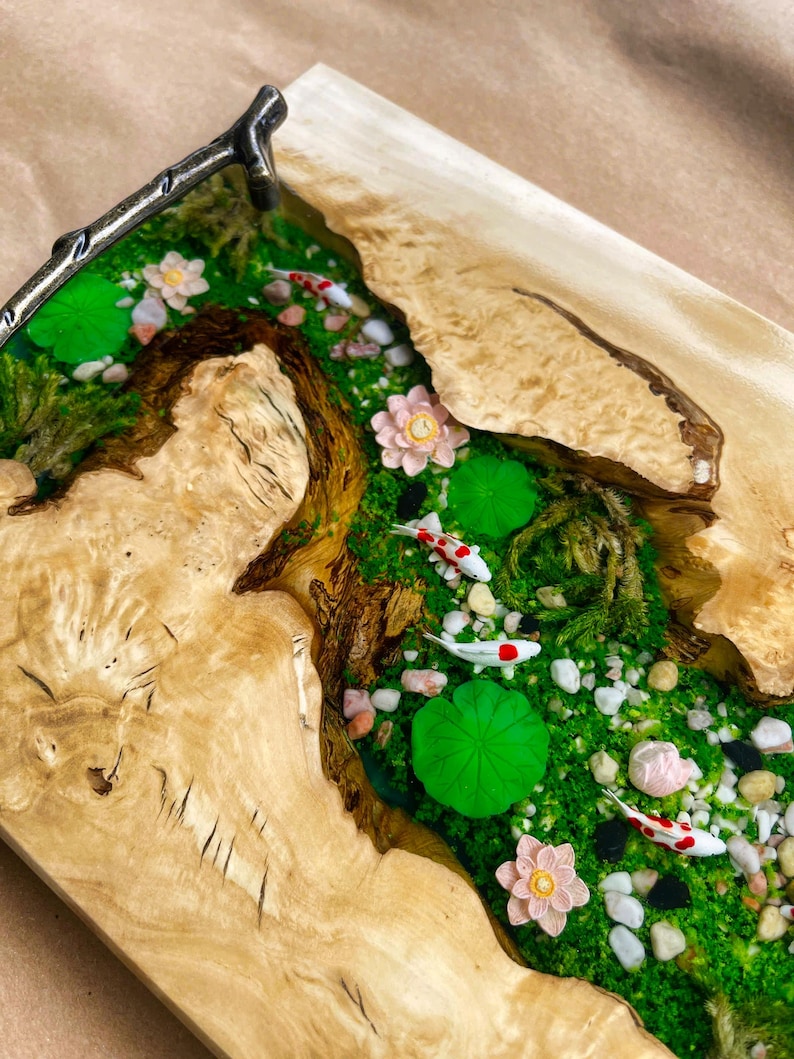 Op de afbeelding: Een houten serveerblad met een riviersc&egrave;ne. De rivier is gevuld met groen mos, wit en rode koi-karpers, roze bloemen en groene waterlelies. De lade heeft een lichtbruine houtnerf.