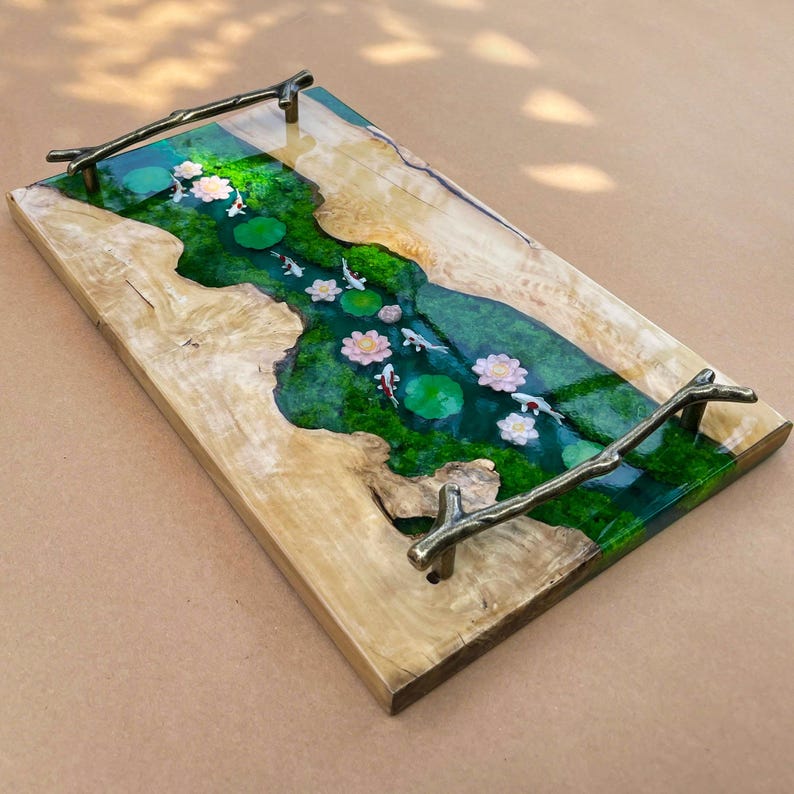 May include: A rectangular wooden serving tray with a river design. The river is filled with green resin, lily pads, koi fish, and pink flowers. The tray has bronze branch handles.
