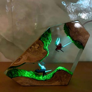 May include: A unique resin art piece featuring two small character figurines within a clear, geometric enclosure. The figures are set against a backdrop of green moss and wood, with a glowing effect. The wooden base has a natural grain.