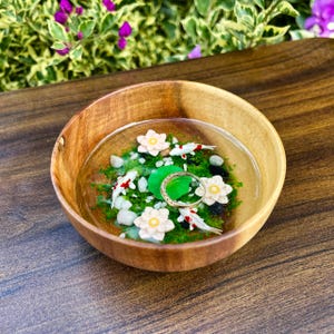 May include: A round wooden bowl containing a clear resin pond scene with green moss, white pebbles, three small white and red koi fish, three white lotus flowers, and a thin gold ring resting on a green lily pad in the center of the bowl.