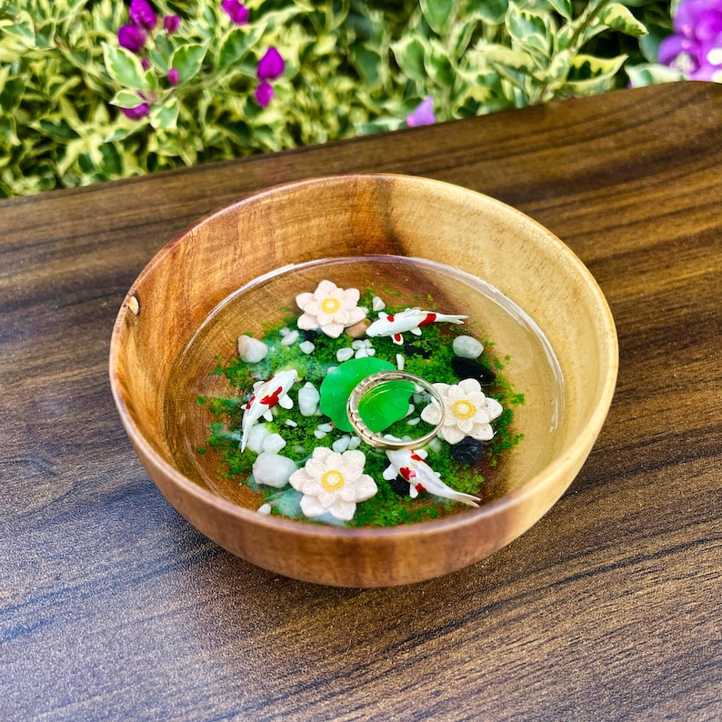 May include: A round wooden bowl containing a clear resin pond scene with green moss, white pebbles, three small white and red koi fish, three white lotus flowers, and a thin gold ring resting on a green lily pad in the center of the bowl.