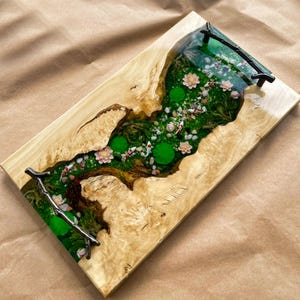 May include: A rectangular wooden serving tray with a river-like design. The centre features a green and brown landscape with miniature koi fish, pink flowers, and green lily pads. The tray has black metal handles.