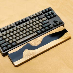 K&ouml;nnte beinhalten: Eine schwarze mechanische Tastatur mit hellgr&uuml;nen Tastenbeschriftungen liegt &uuml;ber einer h&ouml;lzernen Handgelenkauflage. Die Handgelenkauflage hat ein flussartiges Design mit einem dunkelblauen Harzmittelteil und hellen Holzkanten.