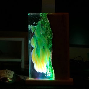 May include: A rectangular resin art piece featuring a vibrant yellow and green betta fish. The fish is suspended in clear resin, with a textured green edge and a dark brown wooden backing. The base is a light-colored wood.