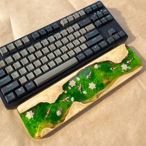 Puede incluir: Un teclado mec&aacute;nico negro con teclas grises se encuentra sobre un reposamu&ntilde;ecas de madera. El reposamu&ntilde;ecas presenta un dise&ntilde;o de r&iacute;o de resina epoxi verde con peces koi, flores blancas y peque&ntilde;as rocas. El teclado tiene un dise&ntilde;o QWERTY est&aacute;ndar.