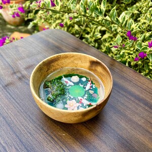 May include: A small wooden bowl containing a clear resin pour that mimics a miniature pond. Inside the resin are three small white and red koi fish, a green lily pad, a pink lotus flower, green moss-like textures, and small white pebbles.