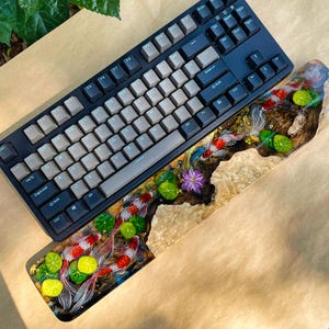 May include: A black mechanical keyboard with gray keycaps sits above a custom wrist rest. The wrist rest features a detailed resin design of koi fish, lily pads, and a purple flower, creating a vibrant and artistic aesthetic.
