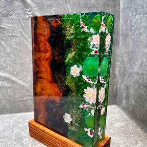 May include: A rectangular resin art piece featuring a koi pond design. The piece has a wooden base and showcases a vibrant scene with koi fish, lotus flowers, and greenery. The left side has a wood grain pattern.