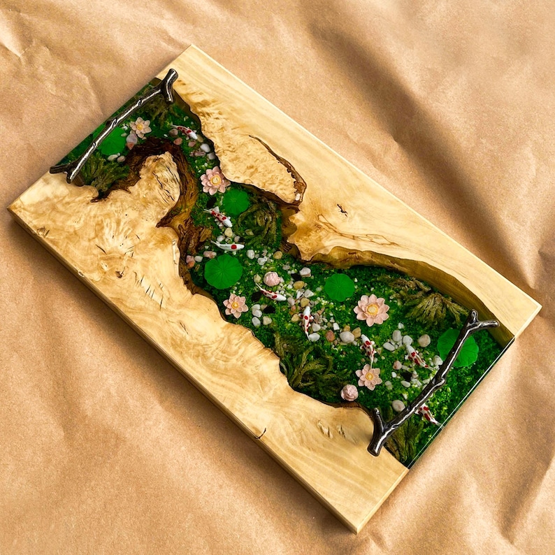 Op de afbeelding: Rechthoekig houten dienblad met een rivierontwerp. De rivier is gevuld met groen mos, waterlelies, kleine vissen en roze bloemen. Het hout heeft een natuurlijke nerf en een lichtbruine kleur.
