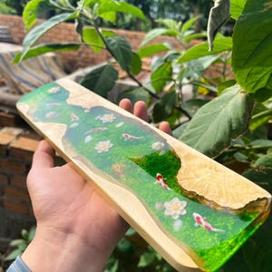Puede incluir: Tabla de servir rectangular de madera con un dise&ntilde;o de r&iacute;o de resina epoxi transparente. La resina presenta una base verde con flores blancas y peces koi rojos y blancos. La tabla tiene un borde de madera natural.