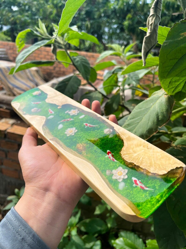 Puede incluir: Tabla de servir rectangular de madera con un dise&ntilde;o de r&iacute;o de resina epoxi transparente. La resina presenta una base verde con flores blancas y peces koi rojos y blancos. La tabla tiene un borde de madera natural.