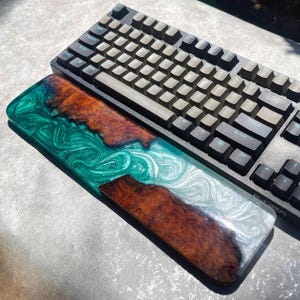May include: A black mechanical keyboard with gray keycaps rests above a custom-made wrist rest. The wrist rest features a unique design with swirling green, white, and brown resin, and a wooden inlay.