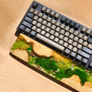 Puede incluir: Un reposamu&ntilde;ecas de madera para teclado con una incrustaci&oacute;n de resina que representa una escena de r&iacute;o y musgo verde con peque&ntilde;os peces blancos y rojos. El reposamu&ntilde;ecas est&aacute; colocado sobre un teclado negro con teclas beige.