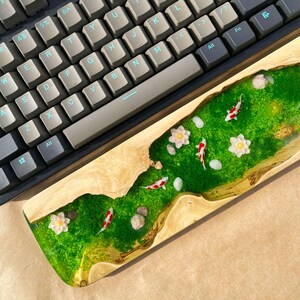Puede incluir: Un reposamu&ntilde;ecas de teclado de madera con un dise&ntilde;o de r&iacute;o de resina epoxi verde. El r&iacute;o presenta peces koi en miniatura, flores blancas y piedras. Un teclado mec&aacute;nico con teclas grises y negras se encuentra encima.