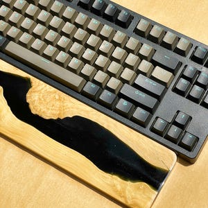 K&ouml;nnte beinhalten: Eine schwarze mechanische Tastatur mit beigefarbenen Tastenkappen und einer h&ouml;lzernen Handgelenkauflage. Die Handgelenkauflage besteht aus hellem Holz mit einem schwarzen Harz-Fluss-Design. Die Tastatur hat ein Standard-QWERTZ-Layout.