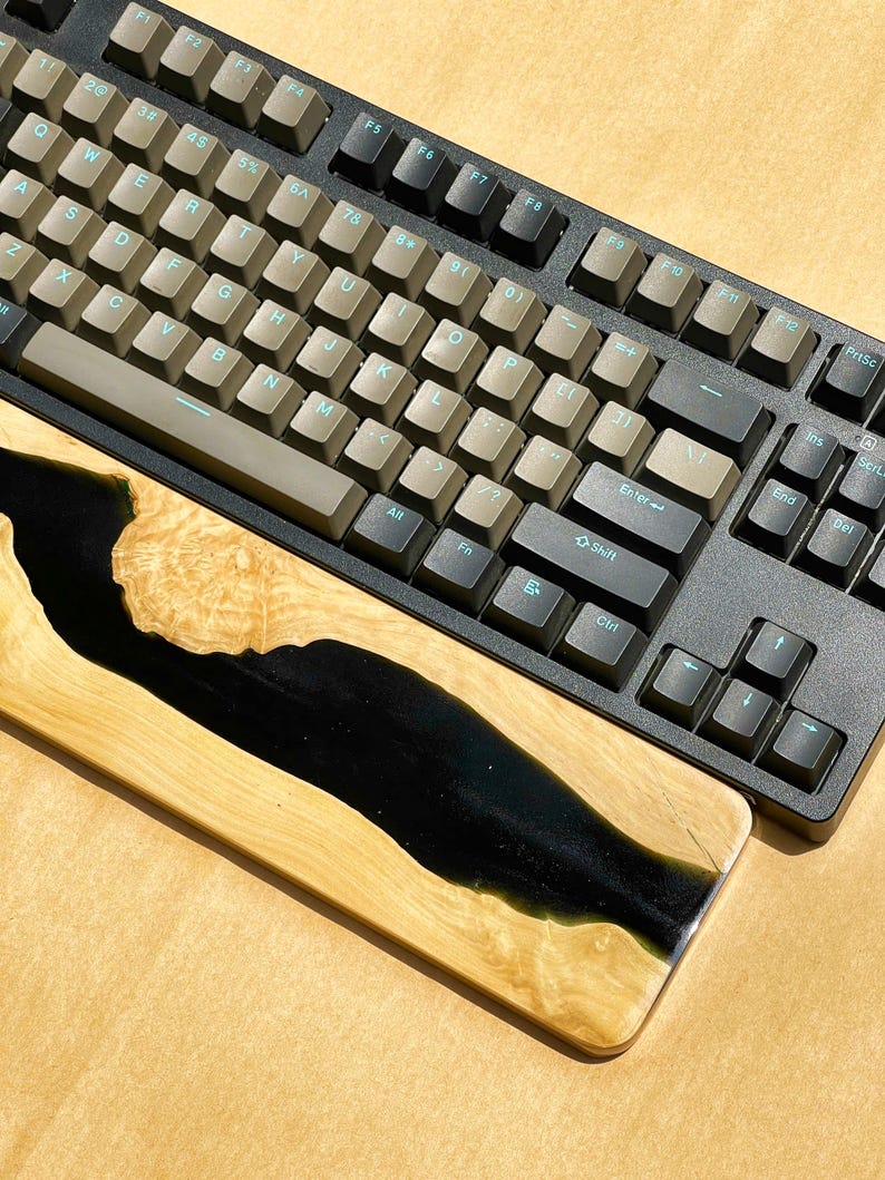 K&ouml;nnte beinhalten: Eine schwarze mechanische Tastatur mit beigefarbenen Tastenkappen und einer h&ouml;lzernen Handgelenkauflage. Die Handgelenkauflage besteht aus hellem Holz mit einem schwarzen Harz-Fluss-Design. Die Tastatur hat ein Standard-QWERTZ-Layout.