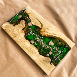 Op de afbeelding: Een rechthoekig houten serveerblad met een rivierachtig ontwerp. Het midden heeft een groen mosachtig landschap met kleine witte en rode vissen, roze bloemen en groene waterlelies. Het hout heeft een lichtbruine kleur.