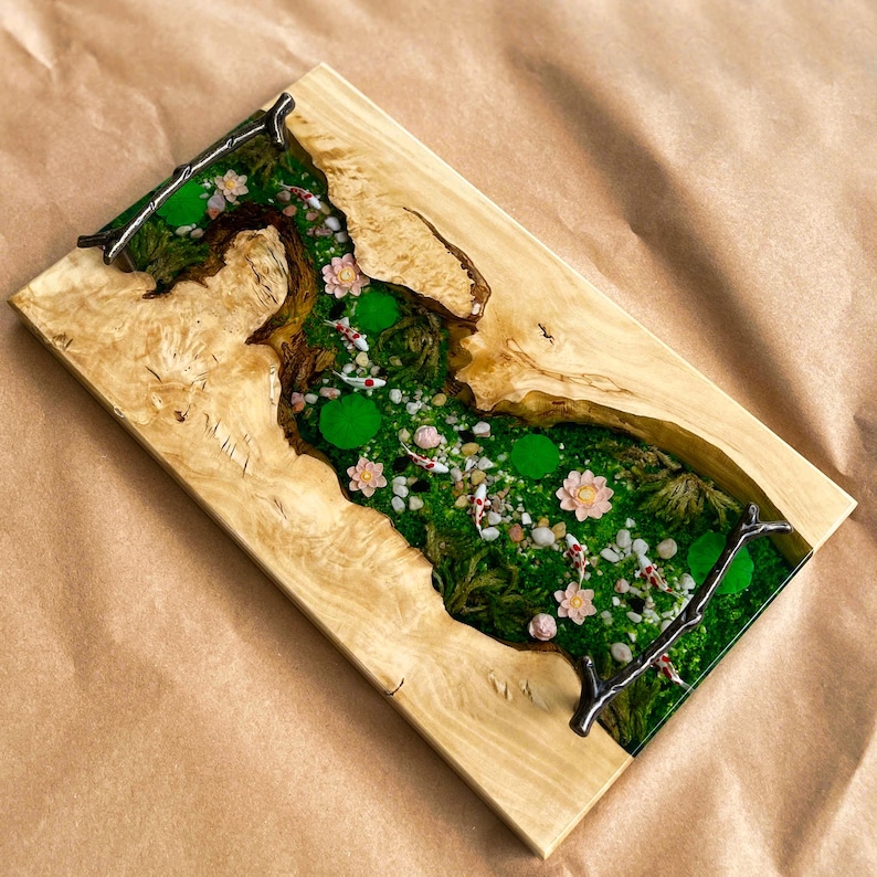 Op de afbeelding: Een rechthoekig houten serveerblad met een rivierachtig ontwerp. Het midden heeft een groen mosachtig landschap met kleine witte en rode vissen, roze bloemen en groene waterlelies. Het hout heeft een lichtbruine kleur.