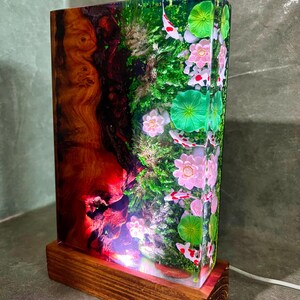 May include: A rectangular resin lamp with a wooden base. The lamp's interior features a vibrant scene with koi fish, pink flowers, and green lily pads. The left side has a red and brown wood grain design. A white cord extends from the base.