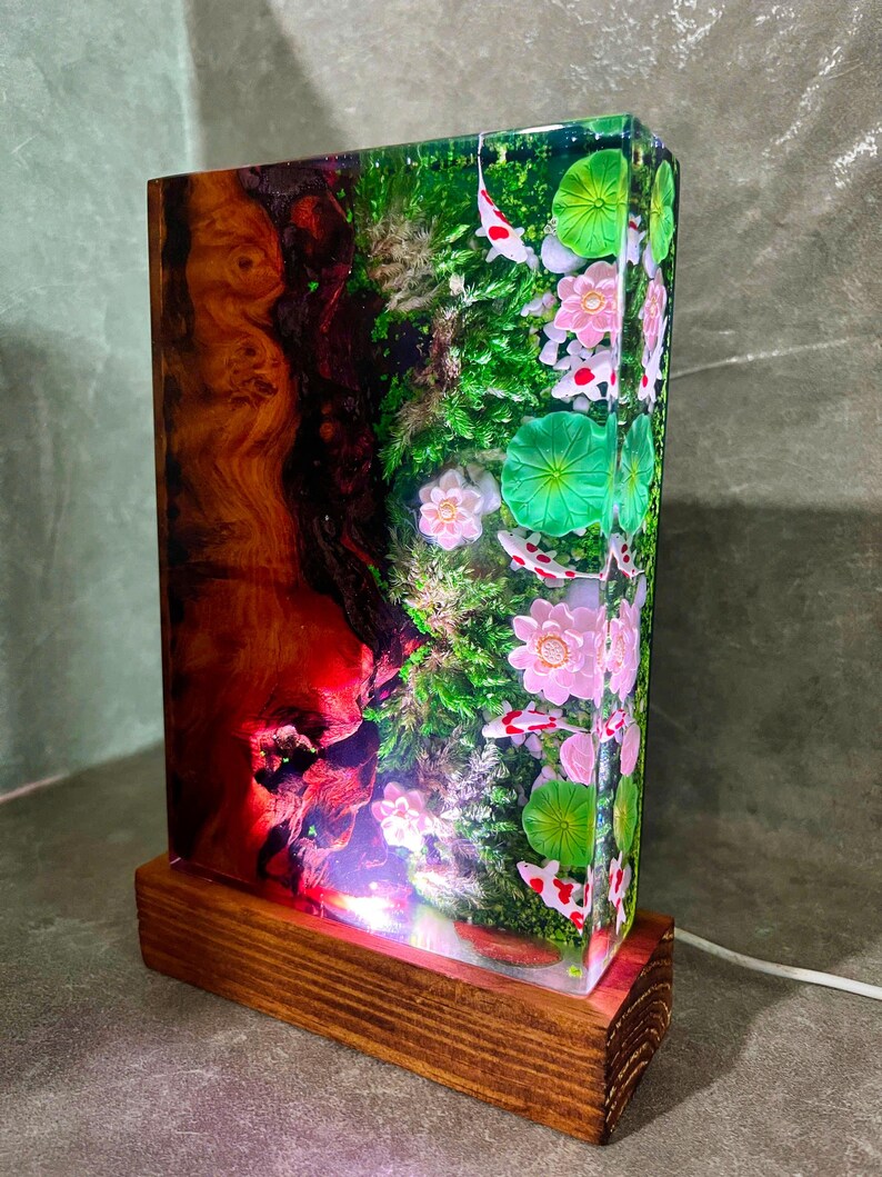 May include: A rectangular resin lamp with a wooden base. The lamp's interior features a vibrant scene with koi fish, pink flowers, and green lily pads. The left side has a red and brown wood grain design. A white cord extends from the base.