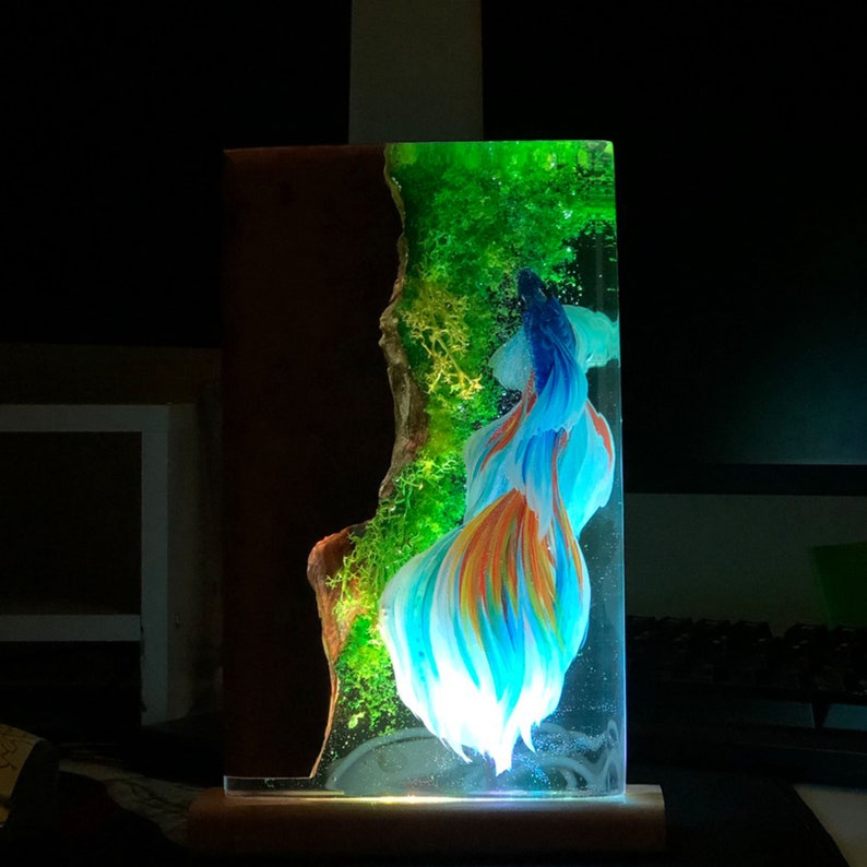 May include: A rectangular resin art piece featuring a vibrant betta fish with blue, orange, and white fins. The artwork includes a brown wooden section and a green, moss-like element, all encased in clear resin. The base is wooden.
