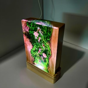 Puede incluir: L&aacute;mpara rectangular de madera con una escena interior detallada. La l&aacute;mpara presenta un vibrante paisaje verde musgoso con peces koi blancos y rojos y flores rosas. La l&aacute;mpara est&aacute; iluminada desde dentro, creando un brillo c&aacute;lido.