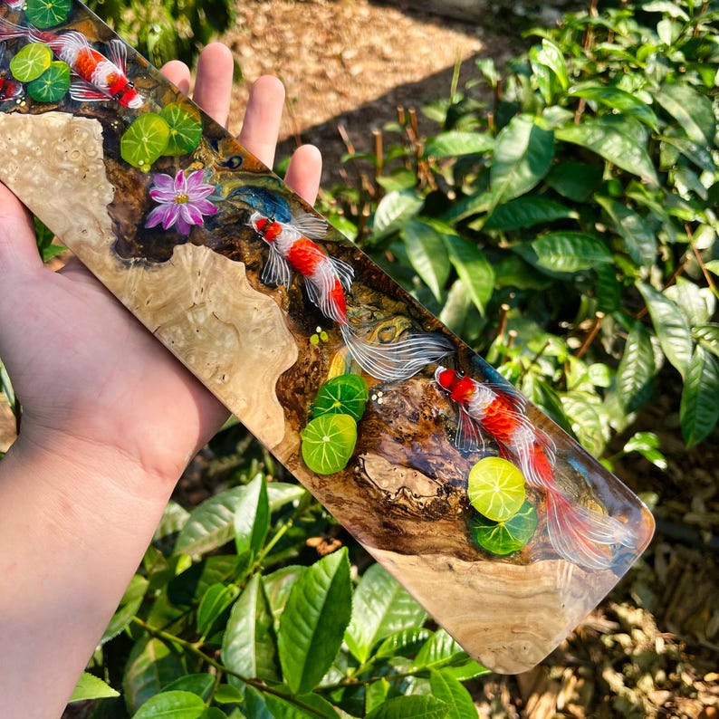 May include: A long, rectangular wooden art piece featuring a koi pond scene. The piece has a natural wood grain base with a clear resin overlay. Red and white koi fish swim among green lily pads and a pink flower.