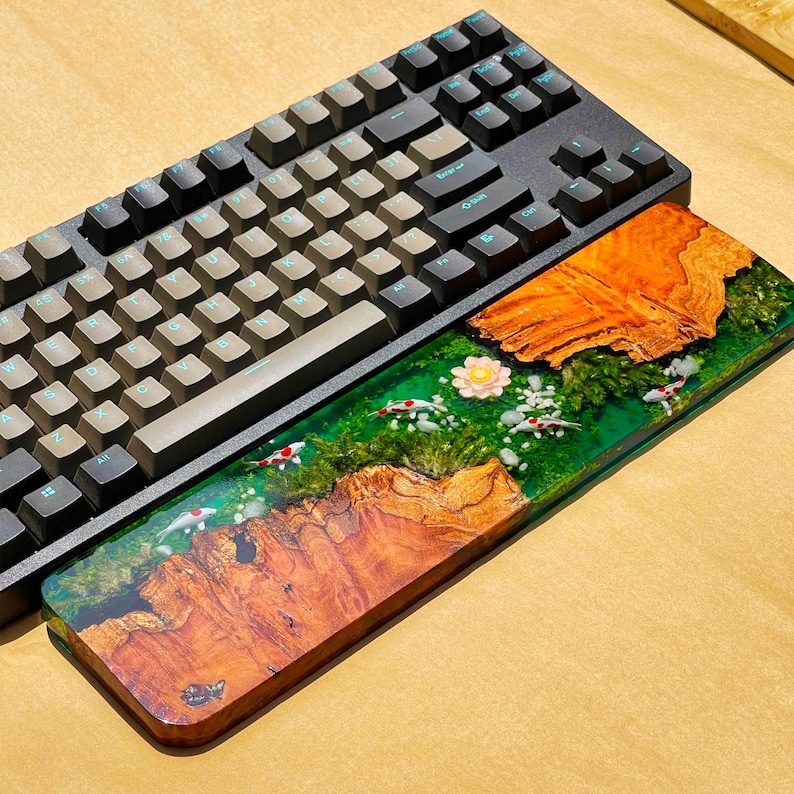 May include: A black mechanical keyboard with gray keycaps rests above a custom wooden wrist rest. The wrist rest features a vibrant design with koi fish, greenery, and a flower set in a clear resin base, with a wood grain pattern.