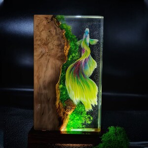May include: A rectangular resin art piece featuring a vibrant betta fish with flowing yellow, green, and purple fins. The fish is set against a backdrop of green moss and a wooden burl, encased in clear resin. The piece is mounted on a dark wooden base.