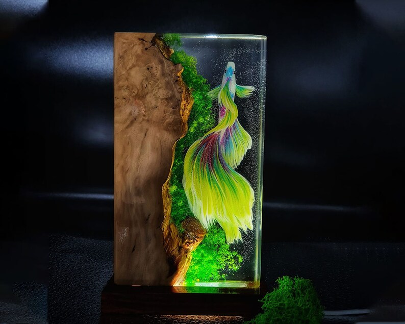 May include: A rectangular resin art piece featuring a vibrant betta fish with flowing yellow, green, and purple fins. The fish is set against a backdrop of green moss and a wooden burl, encased in clear resin. The piece is mounted on a dark wooden base.