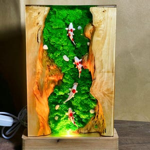 May include: A rectangular wooden lamp featuring a green moss landscape with small white stones and several koi fish with red and white markings. The lamp has a wooden base and a light cord.