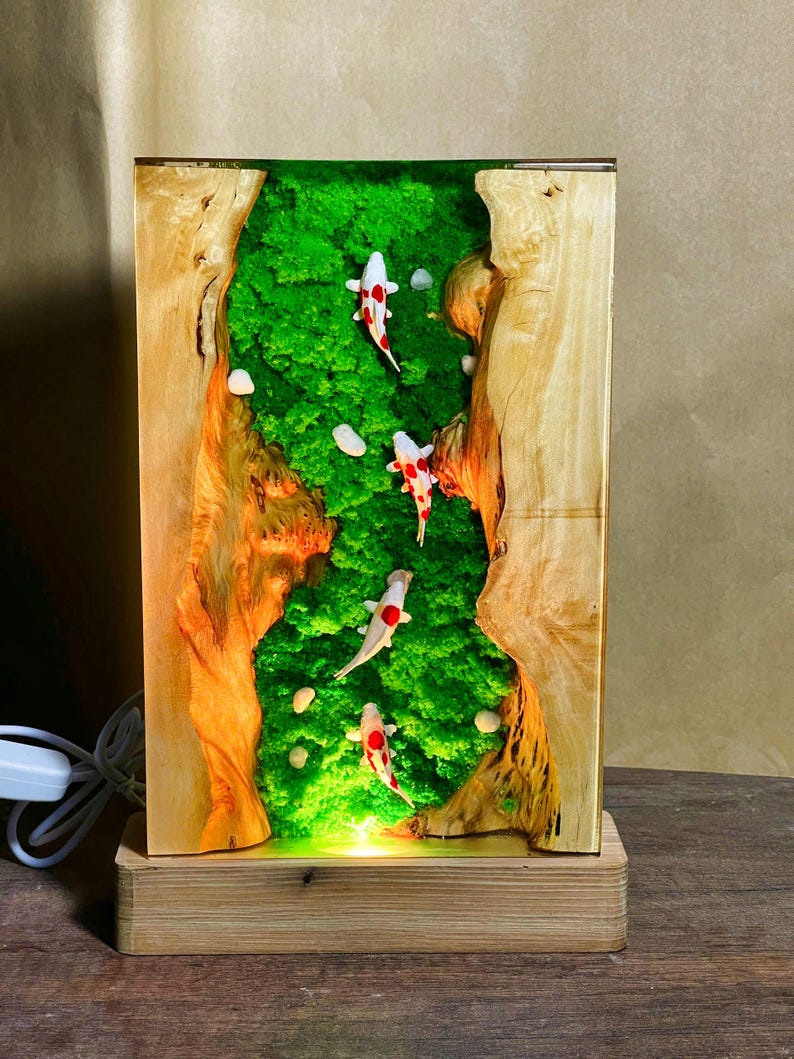 May include: A rectangular wooden lamp featuring a green moss landscape with small white stones and several koi fish with red and white markings. The lamp has a wooden base and a light cord.
