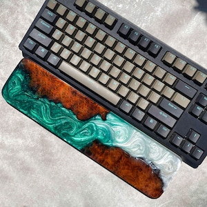 May include: A black mechanical keyboard with beige keycaps rests above a custom resin wrist rest. The wrist rest features a swirling design of green, white, and brown resin, creating a unique aesthetic. The keyboard has a standard layout with function keys.