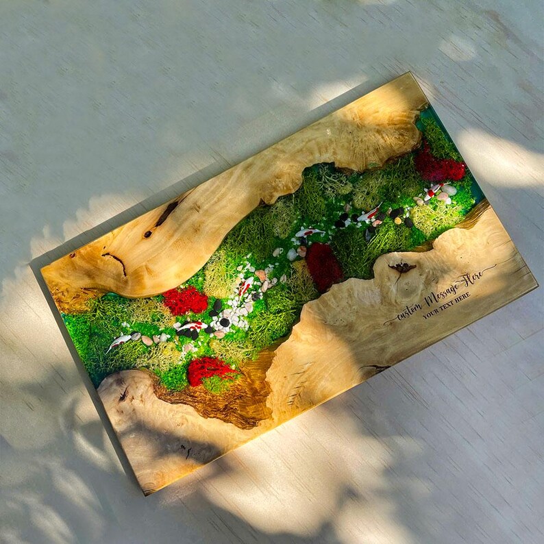 May include: A rectangular wooden serving board with a river-like design. The center features green moss, red accents, and small white and black stones. The edges are light brown wood. Text on the bottom right says "custom message here".