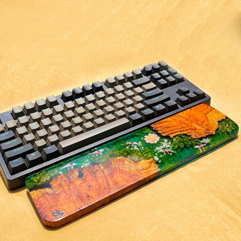 May include: A black mechanical keyboard with gray keycaps rests above a wooden wrist rest. The wrist rest features a vibrant epoxy resin design with a koi pond scene, showcasing orange wood grain, green resin, and white and red koi fish.
