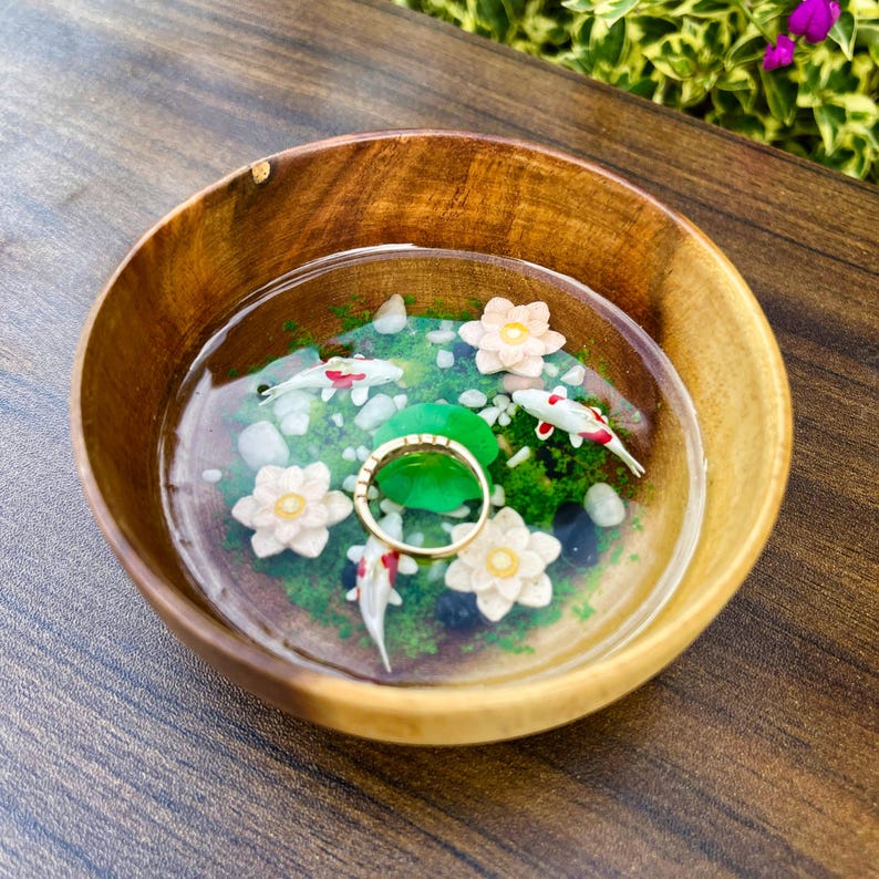 May include: A round wooden bowl containing a clear resin inlay that mimics a pond. The scene features three miniature koi fish in white and red, three white lotus flowers, green moss-like accents, and a gold-toned ring resting in the center.