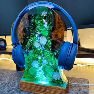 May include: A unique lamp featuring a resin design with koi fish, water lilies, and green foliage. The lamp is mounted on a wooden base and has blue headphones resting on top. The lamp is illuminated with a soft, green light.