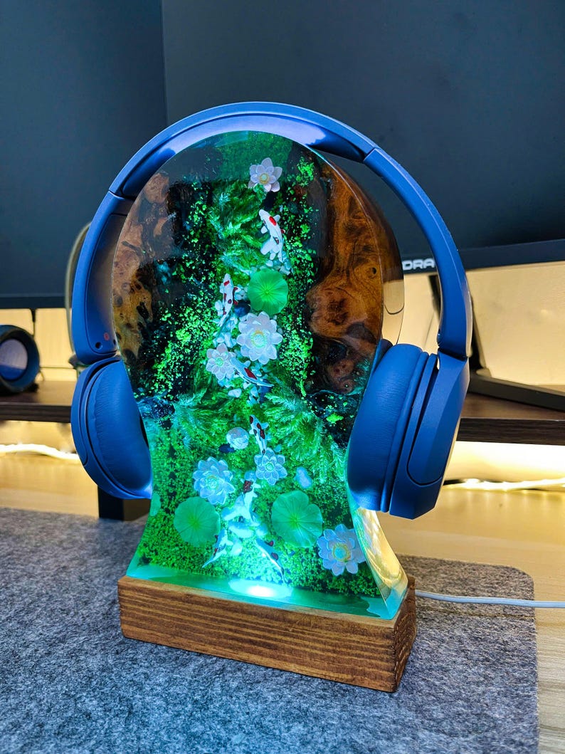 May include: A unique lamp featuring a resin design with koi fish, water lilies, and green foliage. The lamp is mounted on a wooden base and has blue headphones resting on top. The lamp is illuminated with a soft, green light.