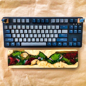 May include: A black mechanical keyboard with a white keycap set and a wooden keyboard rest with a green and red resin design. The keyboard rest has a river scene with fish and plants.