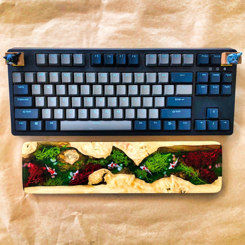 May include: A black mechanical keyboard with a white keycap set and a wooden keyboard rest with a green and red resin design. The keyboard rest has a river scene with fish and plants.