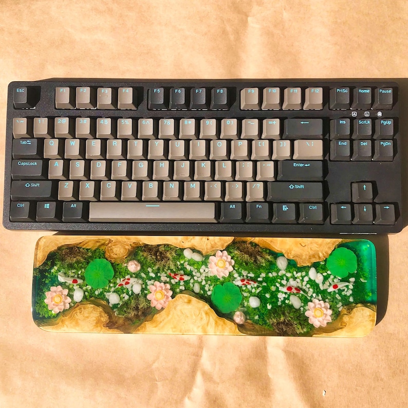 Pu&ograve; includere: Una tastiera meccanica nera con un set di tasti marroni e un poggiapolsi in legno personalizzato con un'intarsio in resina verde, bianca e rosa. Il poggiapolsi presenta un design a forma di fiume con fiori e foglie.