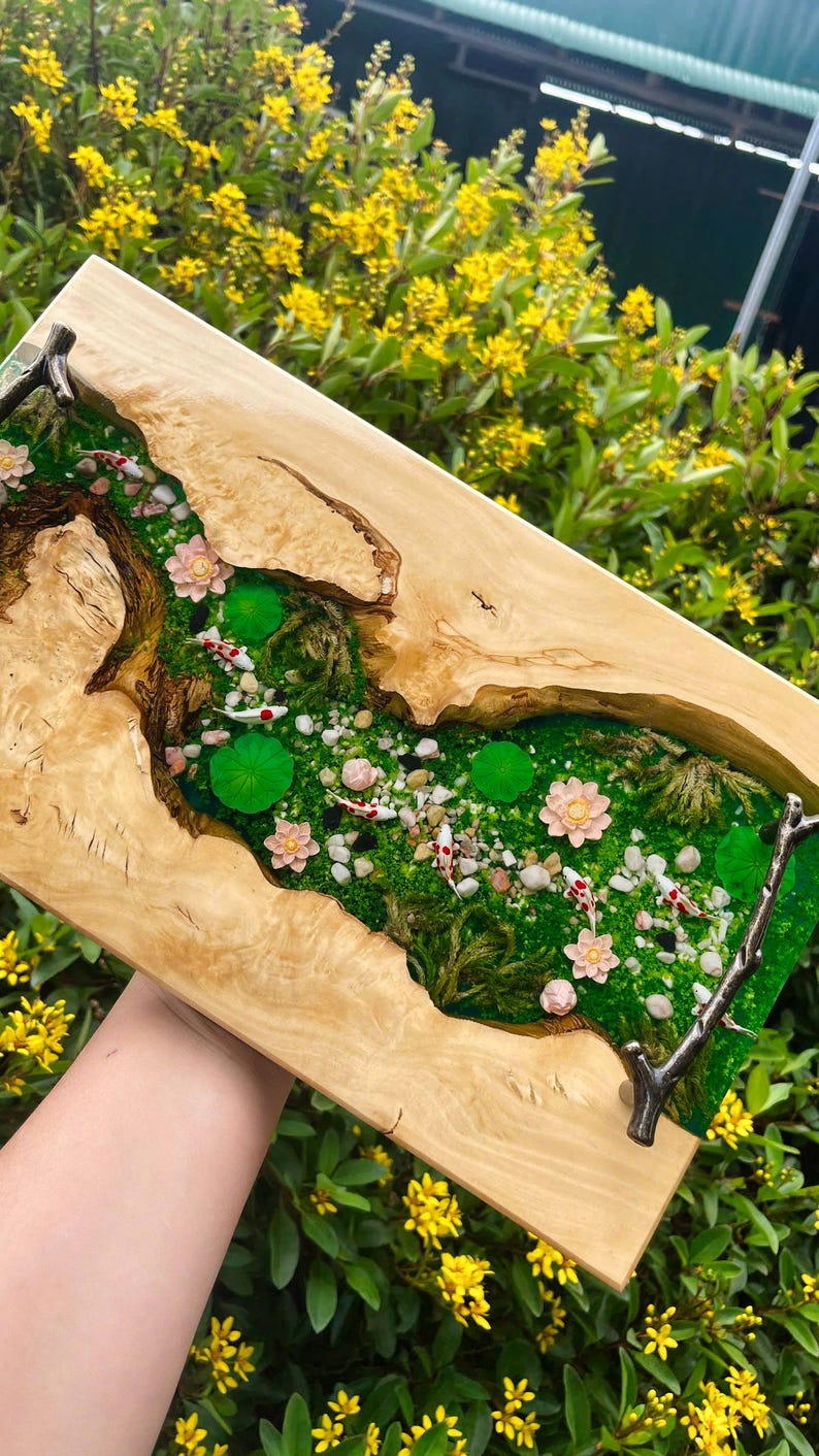 Op de afbeelding: Een rechthoekig houten kunstwerk met een miniatuurvijver. De vijver is gevuld met groen mos, kleine steentjes, waterlelies en roze bloemen. Verschillende koikarpers zwemmen in het water. Het houten frame heeft een natuurlijke houtnerf.