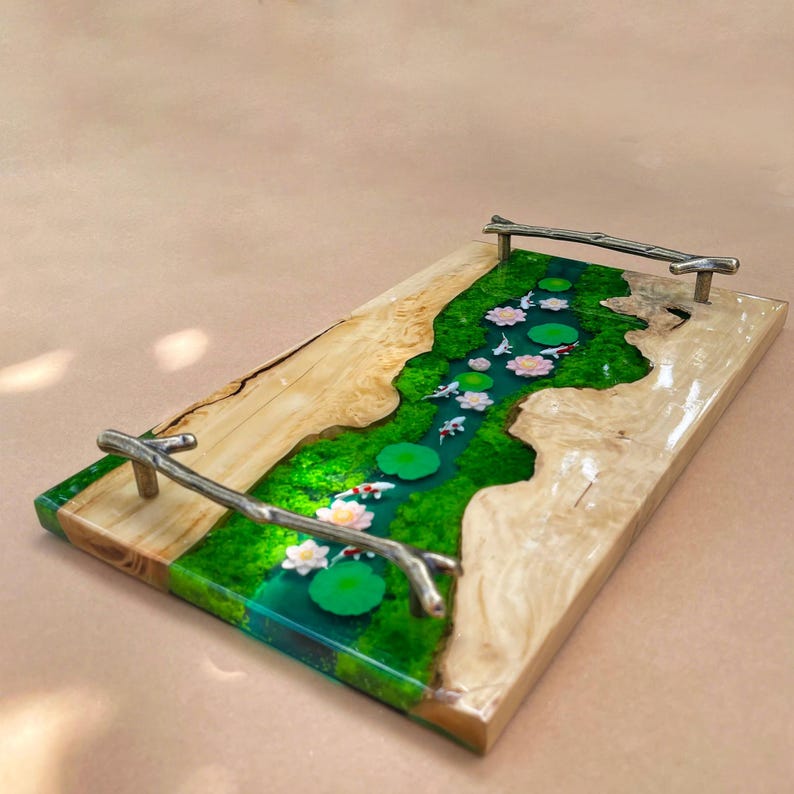 May include: A rectangular wooden serving tray with a river design. The river is made of green resin with white and pink lotus flowers, lily pads, and koi fish. The tray has silver branch-shaped handles.