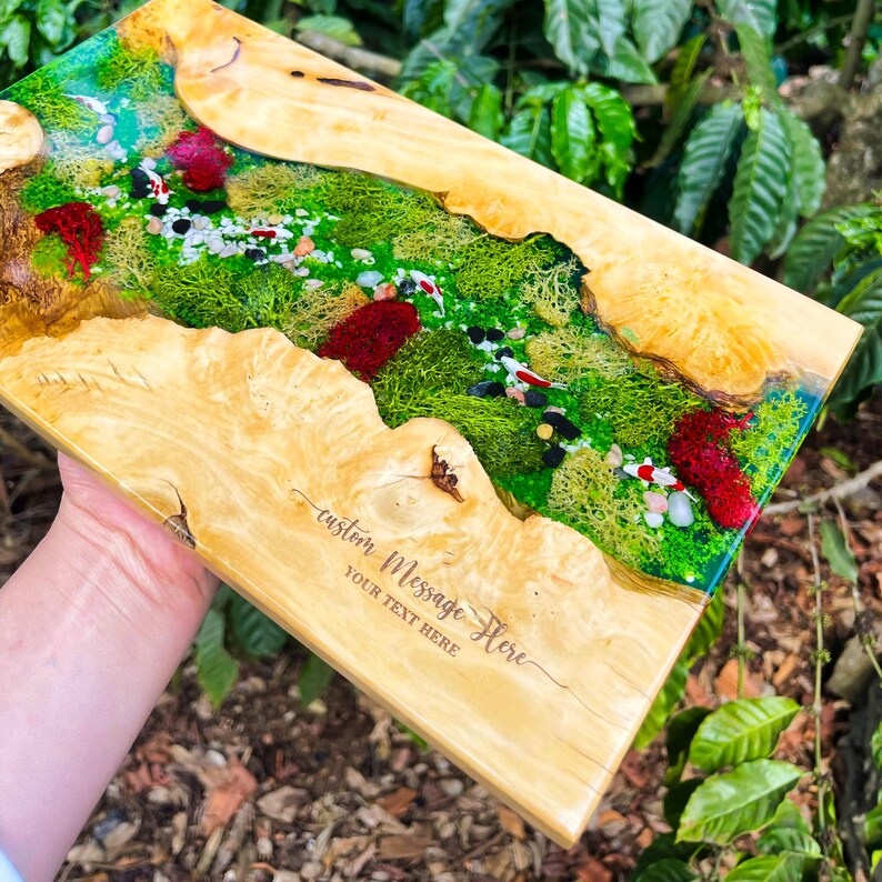 May include: A rectangular wooden serving board with a river scene design. The river features green moss, small stones, and red accents, encased in clear resin. The board has the text "custom Message Here" and "YOUR TEXT HERE" engraved on the wood.
